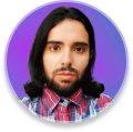 Gabs with a serious look, wearing a red plaid shirt, beard, shoulder-length black hair, lilac gradient background.